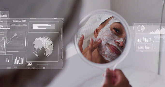 Image of graphs, globes over biracial woman looking in mirror and applying cream on face