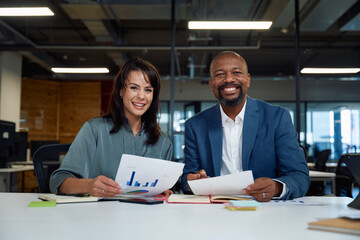 Two happy multiracial colleagues looking at camera and smiling with graph documents in office