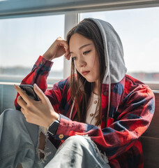 Beautiful serious Asian teenage girl sitting by the window and looking at the smartphone screen