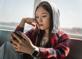 Beautiful serious Asian teenage girl sitting by the window and looking at the smartphone screen