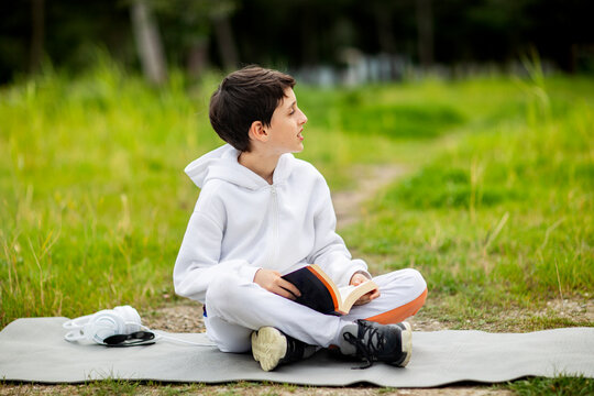 cute boy sitting with book on yoga mat against forest bacground