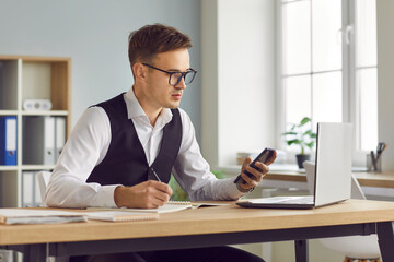 Confident young business man in suit writing tasks and making notes in notepad sitting at the desk on workplace with laptop with smartphone in hand at office planning daily appointment.