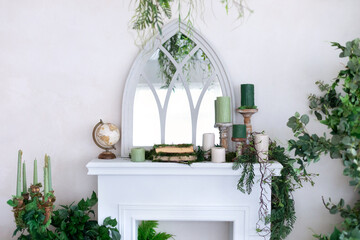 Fireplace with a mirror above it. Interior design living room with a white fireplace and carved mirror. Fireplace decorated with candles, stabilization moss, decor, globe, green plants and books.	

