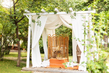 Summer gazebo with flowing white curtains. Wedding boho decoration. Decor outdoor terrace with wicker furniture. Outdoor design of arbour. Rattan peacock armchair and decorative folding screen divider © stock_studio