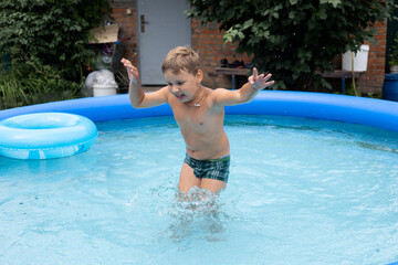 Naklejka premium In the summer, a boy child with his eyes closed jumps into the water in the pool and raises a lot of splashes. A child loves to swim in the pool in the yard of his house on a hot summer day.