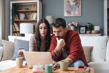 Young couple making shopping online with credit card and laptop at home. Two happy people holding debit card while buying on e-commerce site using laptop, or making reservation or holidays. - Powered by Adobe