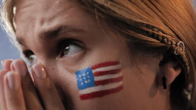 USA sports fan is worried. Close-up of face of young female fan with US flag on her face, woman watches match, competition, game and is worried, cheers for USA