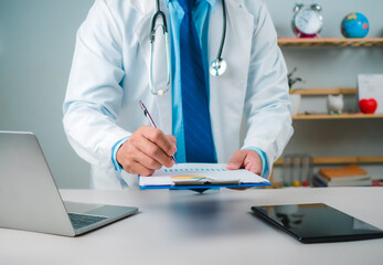 Male doctor sitting at work desk checking work plans and writing document reports in hospital office, healthcare worker and doctor service, digital laptop in modern office, medical technology concept.