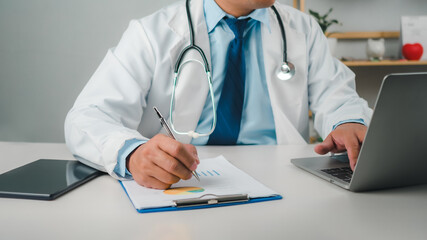 Male doctor sitting at work desk checking work plans and writing document reports in hospital office, healthcare worker and doctor service, digital laptop in modern office, medical technology concept.