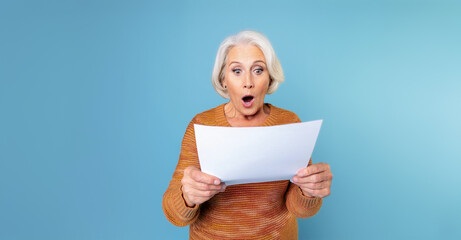 Senior lady with gray hair, shocked expression and mouth open, reading a white piece of paper in her hands. Isolated on blue background.