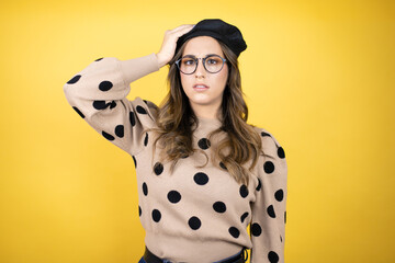 Young beautiful brunette woman wearing french beret and glasses over yellow background putting one...