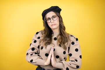 Young beautiful brunette woman wearing french beret and glasses over yellow background begging and...