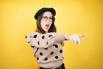 Young beautiful brunette woman wearing french beret and glasses over yellow background pointing...