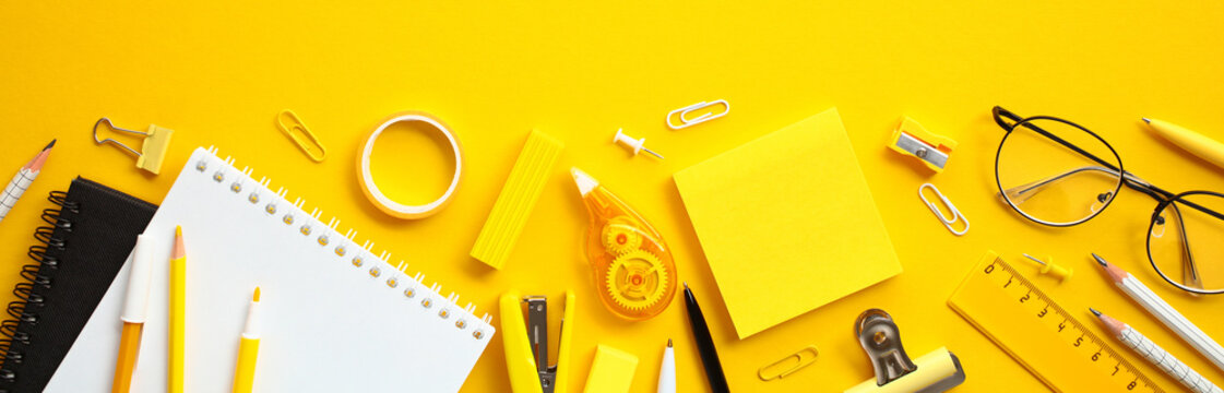 Flat lay office supplies on yellow background. Wide banner with paper notebook, ruler, pens, pencils, eyeglasses, sticky notes and other stationery.
