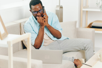 Smiling African American Man using Laptop and Phone on Sofa in Home Office for Online Shopping and...