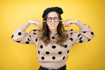Young beautiful brunette woman wearing french beret and glasses over yellow background covering...