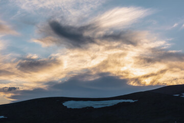 Magnificent sunset cloudy glowing sky of gold colors. Mountain silhouette with snow under dawn skies of sunrise vivid orange tones. Beautiful contrast texture of golden clouds in colorful luminous sky