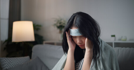 Portrait of Unwell young woman have a high fever and massaging temples to relieve headache ,wrapped in blanket sitting on sofa in the living room at home, healthcare woes