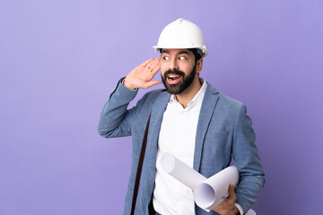 Young architect man with helmet and holding blueprints over isolated purple background listening to something by putting hand on the ear