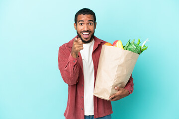 Young latin man holding a grocery shopping bag isolated on blue background surprised and pointing...