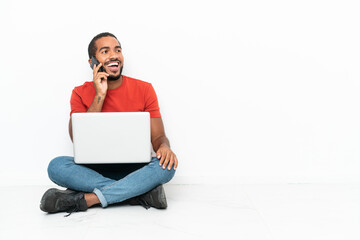 Young Ecuadorian man with a laptop sitting on the floor isolated on white background keeping a conversation with the mobile phone