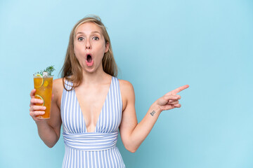 Young blonde woman wearing swimsuit holding a cocktail isolated on blue background surprised and...