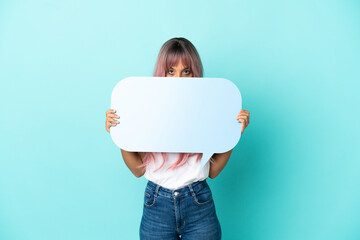 Young mixed race woman with pink hair isolated on blue background holding an empty speech bubble...