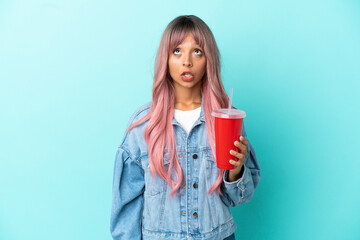 Young mixed race woman drinking a fresh drink isolated on blue background looking up and with surprised expression