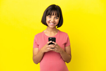 Young mixed race pregnant woman isolated on yellow background looking at the camera and smiling while using the mobile