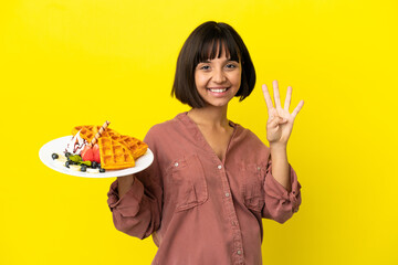 Pregnant woman holding waffles isolated on yellow background happy and counting four with fingers