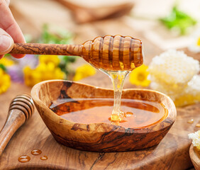 Honey dripping from wooden spoon into wooden bowl with honey, spring flowers are on wooden table...