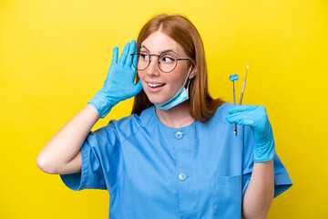 Young redhead Dentist woman isolated on yellow background listening to something by putting hand on the ear