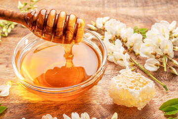 Honey dripping from wooden spoon in glass bowl with organic honey, honey plant acacia flowers and...