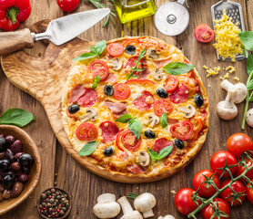 Pizza topped with cherry tomatoes, salami and other ingredients is on wooden table surrounded by ingredients. Top view.