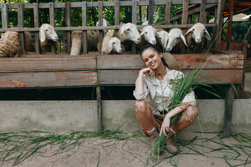 A woman kneeling in front of a fence with sheep in the background, holding a bunch of grass - Powered by Adobe