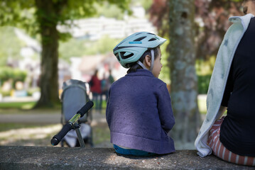 Boy wearing a bike helmet sitting on a park bench with his mother and a scooter nearby, enjoying a...