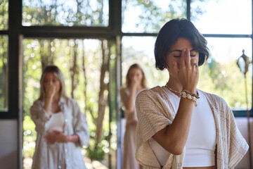 Women group meditating with eyes closed doing yoga pranayama breathing exercises holding fingers on face feeling energy during holistic healing yoga training class retreat session.