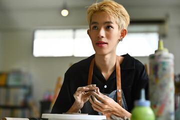 A young LGBT boy with short blonde hair engaged in crafting pottery, The person appears focused and...