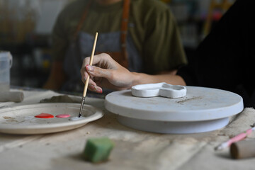 A close-up of an artist's hand holding a brush and mixing paints on a palette. A heart-shaped ceramic piece is being painted on a rotating platform