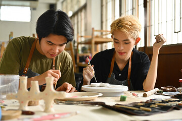 Two students concentrate on painting ceramic pieces in an art studio. They work side by side,...