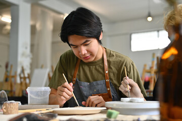Two teenage Asian students focused on painting ceramic pieces in an art classroom together