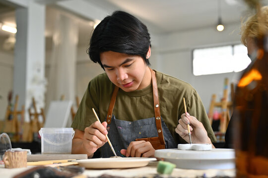 Two teenage Asian students focused on painting ceramic pieces in an art classroom together