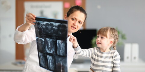 Portrait of smart female doctor sitting in hospital office and showing little lady photo of body condition. Small kid looking with interest and calmness. Health checkup concept