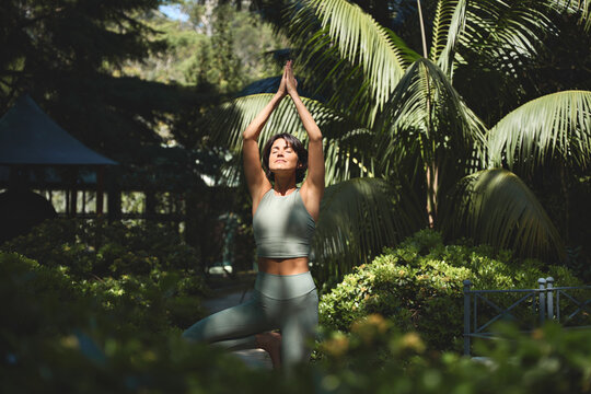 Fit sporty mindful young Hispanic woman meditating doing yoga breathing exercises with eyes closed feeling peace of mind, mental balance standing in green nature tropical park on sunny day.