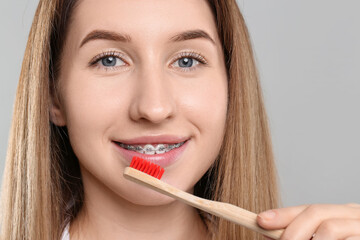 Smiling woman with dental braces cleaning teeth on grey background, closeup