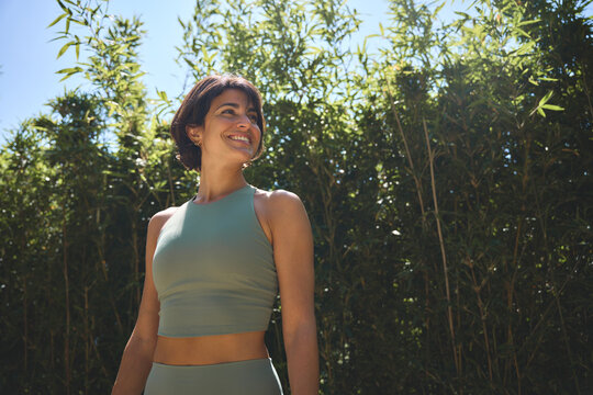 Happy fit sporty active confident young Hispanic woman wearing sportswear enjoying summer sunny day looking away feeling peace of mind, mental balance standing in green nature park.