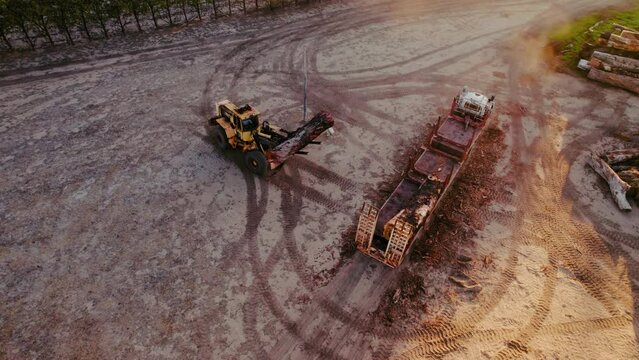 A fork lifter is seen unloading large tree trunks from a truck in a rural, dusty area during sunset, creating circular tire tracks on the ground.