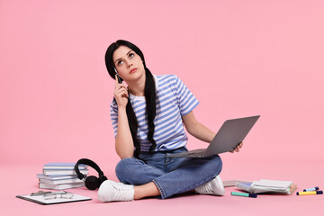 Student with laptop sitting among books and stationery on pink background