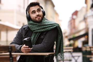 Smiling man in warm scarf with paper cup listening to music in outdoor cafe