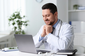 Smiling doctor having online consultation via laptop at table in clinic